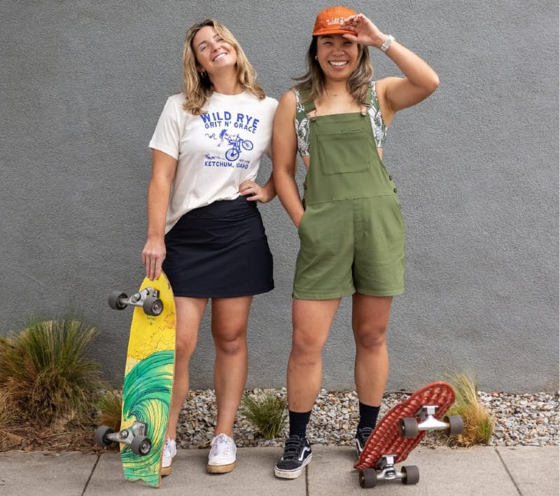 Two women standing with skateboards wearing Wild Rye casualwear—a t-shirt and skirt on one, and overalls on the other.