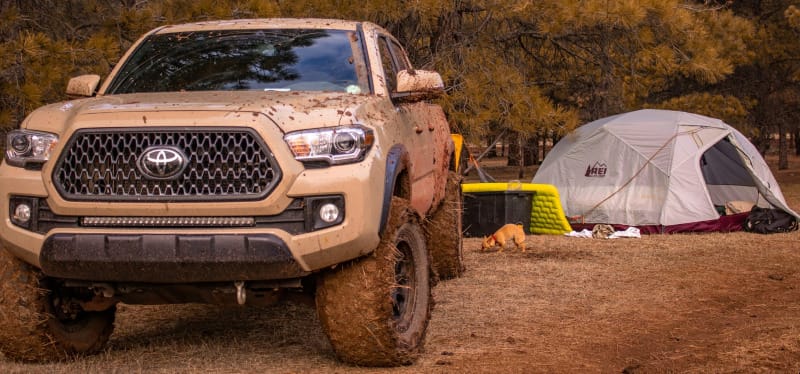 Toyota Tacoma parked at a forest campsite with an REI tent and gear setup in the background.