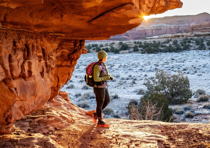 Hiker wearing cold-weather hiking layers and backpack, standing under a red rock alcove with frosty desert landscape in the background, autumn adventure.