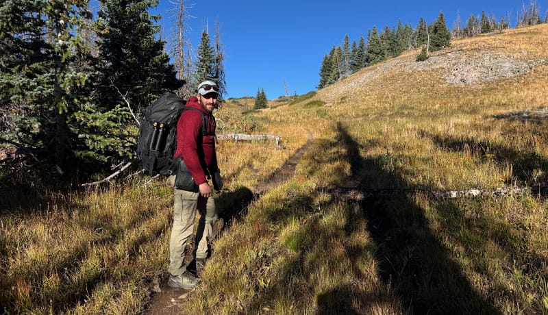 Hiker wearing a red FarPointe Alpha Cruiser midlayer while carrying a large pack on a mountain trail in golden autumn grass.