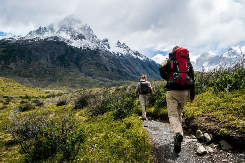 Two hikers with backpacks trekking a mountain trail with snow-covered peaks ahead.