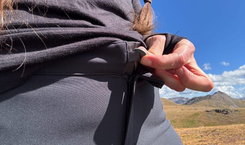 Close-up of a hiker tightening the waistband adjustment on the KUIU Attack Pants against a backdrop of mountain peaks.