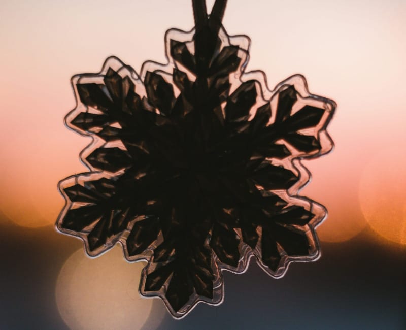 Close-up of a snowflake ornament silhouetted against a warm sunset with bokeh lights.
