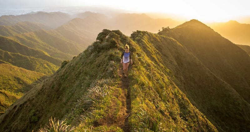 Woman hiking a narrow mountain ridge at golden sunset with layers of mountains in the distance.