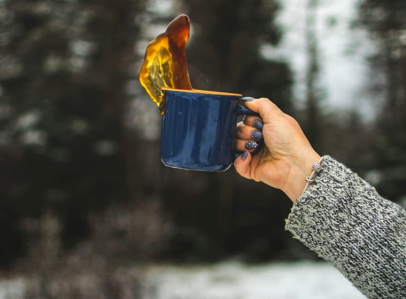 A hand holding a blue enamel camping mug as coffee splashes out against a snowy forest backdrop.