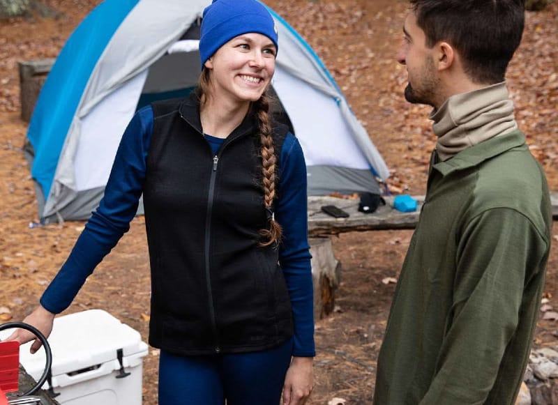 Smiling campers wearing Minus33 merino wool base layers and fleece vest near a tent and camping stove in autumn woods.