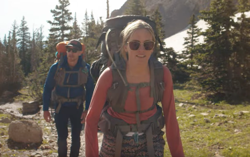 Two hikers wearing Mystery Ranch Bridger 55 backpacks walking through alpine forest with snow patches nearby.