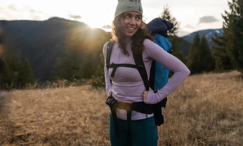 Backpacker wearing lilac Ibex Woolies Pro Tech Half Zip merino base layer with pack and beanie in alpine meadow, illustrating hiking performance and comfort.