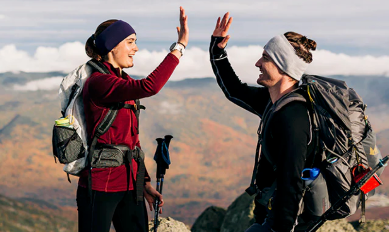 Two hikers wearing Minus33 merino wool layers high-fiving on a mountain summit with autumn scenery in the background.