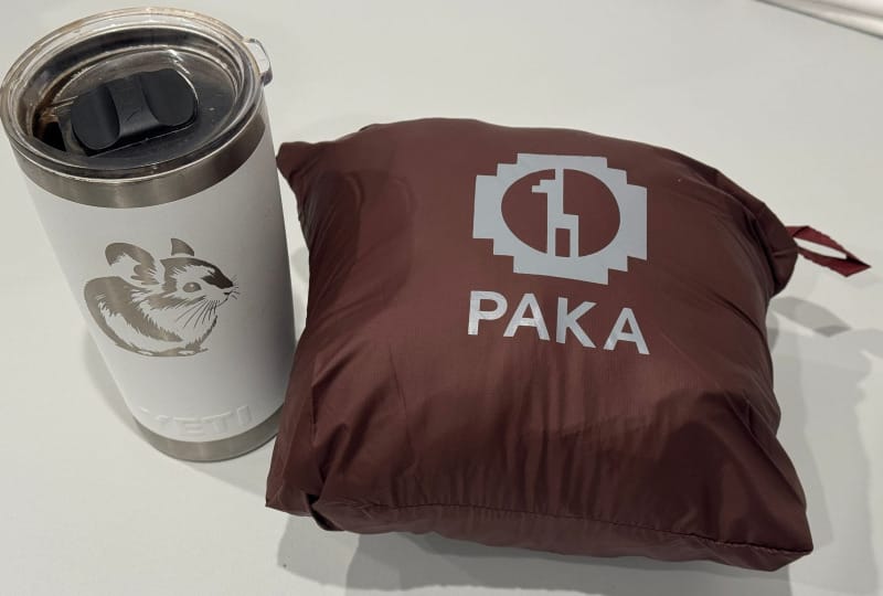 PAKA Mayu Ultralight Jacket packed into stuff sack next to white YETI mug with pika illustration.