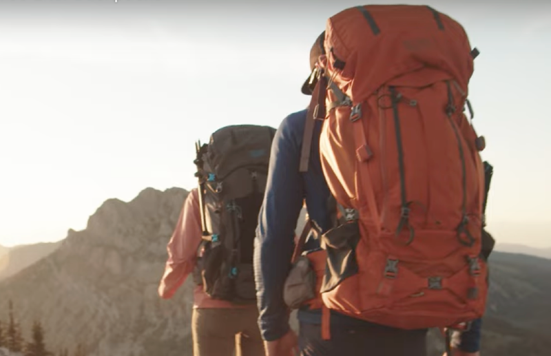 Two hikers wearing Mystery Ranch Bridger 55 backpacks on a mountain trail during golden hour with peaks in the background.