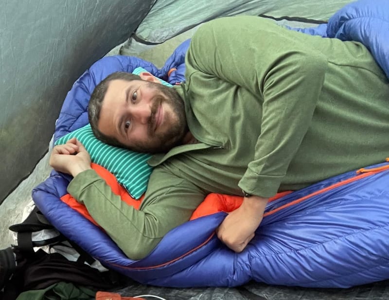 Two backpackers relaxing inside a tent with sleeping bags and gear, one using the NEMO Fillo inflatable pillow for comfort during a camping trip.