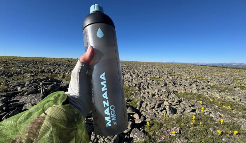 hand holding mazama water bottle on hike in alpine with wildflowers and blue sky