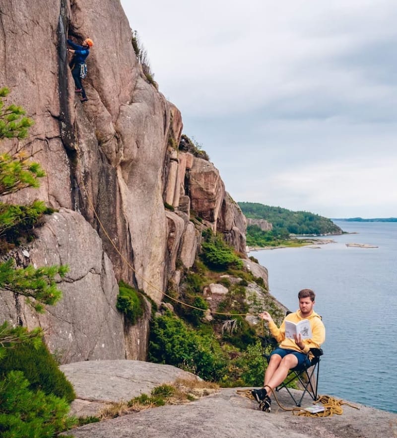 A rock climber ascending a cliff while their belayer sits in a camp chair reading a book overlooking a coastal landscape.