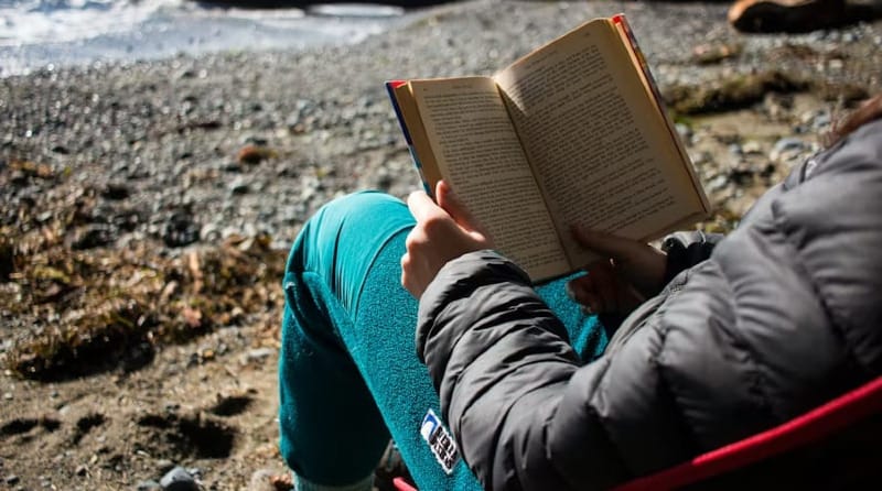 Close-up of someone sitting by a rocky shoreline reading a book with sunlight reflecting off the water.
