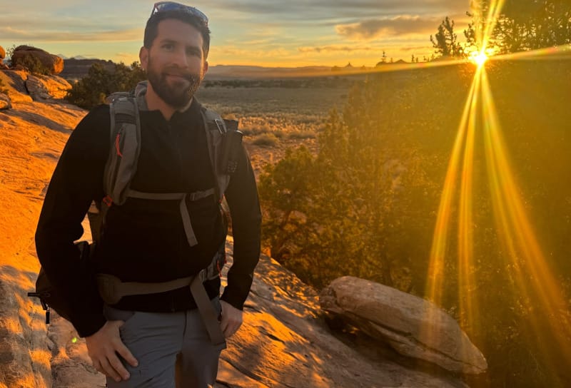 Hiker standing on slickrock at sunset with a backpack, golden sunlight shining through trees and desert terrain behind him.