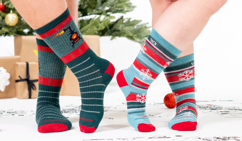 Two people wearing Farm To Feet holiday socks with snowflake patterns beside a Christmas tree and wrapped gifts.