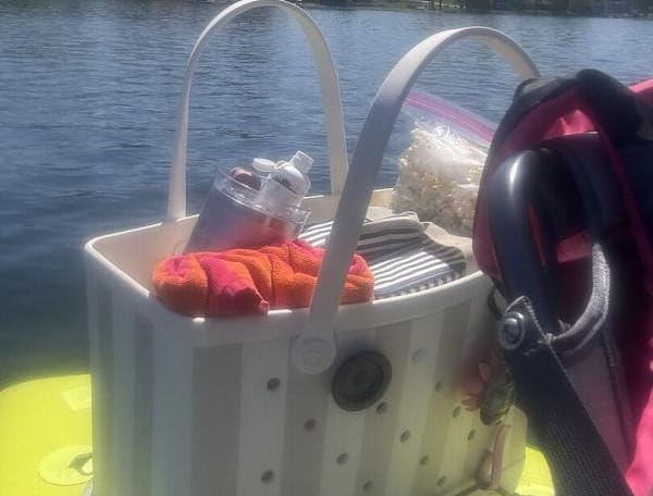 A white Original Bogg Bag filled with outdoor essentials sitting on a paddleboard on a sunny lake, with houses and trees in the background.