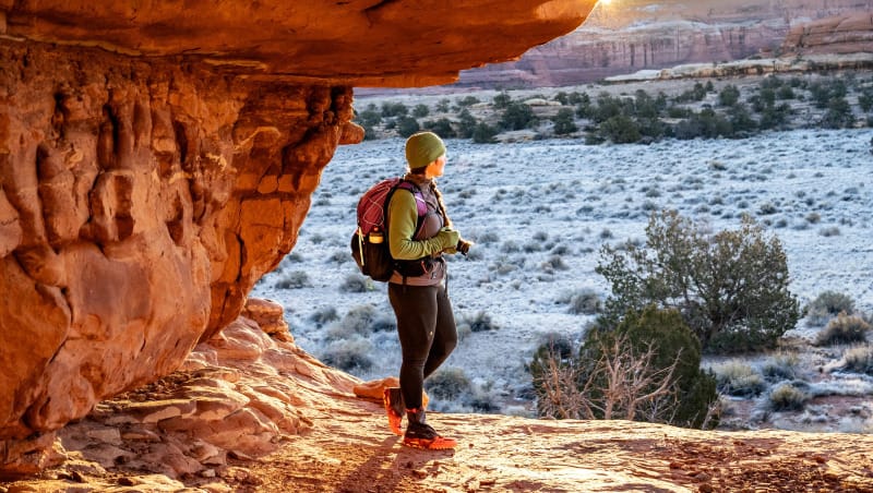 Backpacker standing under a red rock arch at sunrise with snowy desert landscape in the background, wearing winter hiking gear and pack.