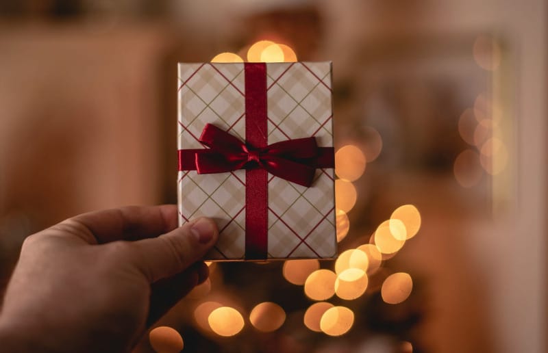 Hand holding a wrapped holiday gift box with a red ribbon in front of warm, out-of-focus Christmas lights.
