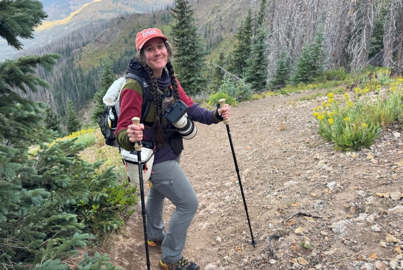Hiker standing on a mountain trail holding Gossamer Gear LT5 carbon trekking poles, surrounded by pines and high-elevation scenery.