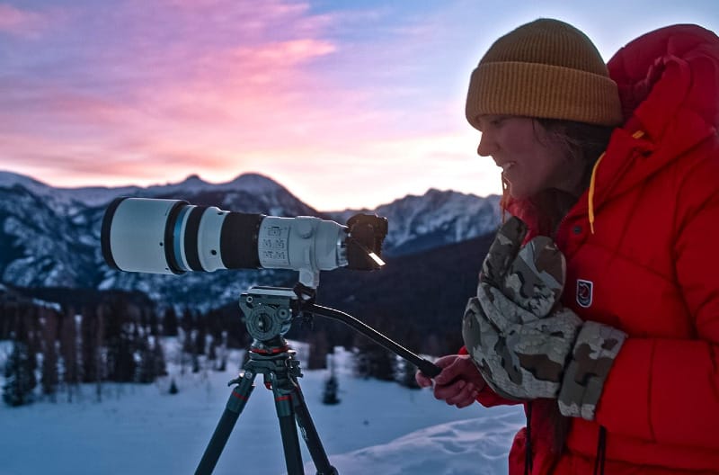 A person wearing KUIU Super Down Pro Glassing Glomitts and a red puffy jacket operates a large telephoto camera setup on a tripod in snowy mountains at sunrise.