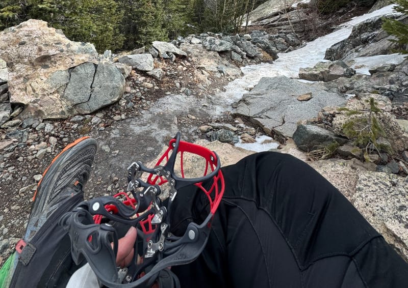 Hiker sitting on a rocky mountain trail holding Kahtoola MICROspikes Ghost traction devices, with steep granite cliffs and patches of ice in the background.