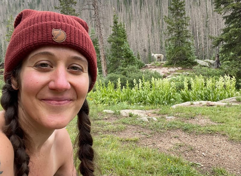 Hiker wearing a red Fjällräven Tab Hat while sitting outdoors in a lush alpine landscape with trees and mountains in the background.