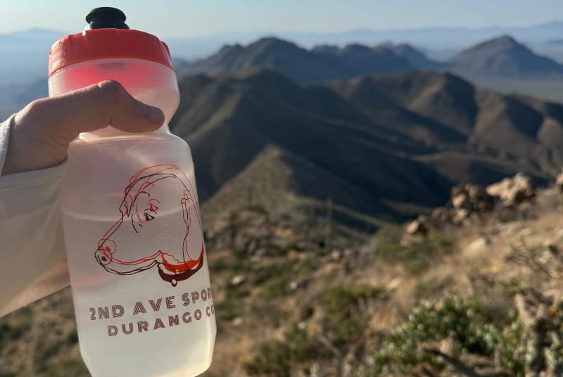 Hand holding a water bottle of electrolytes on a desert mountain hike in the Southwest with hazy ridgelines in the distance.