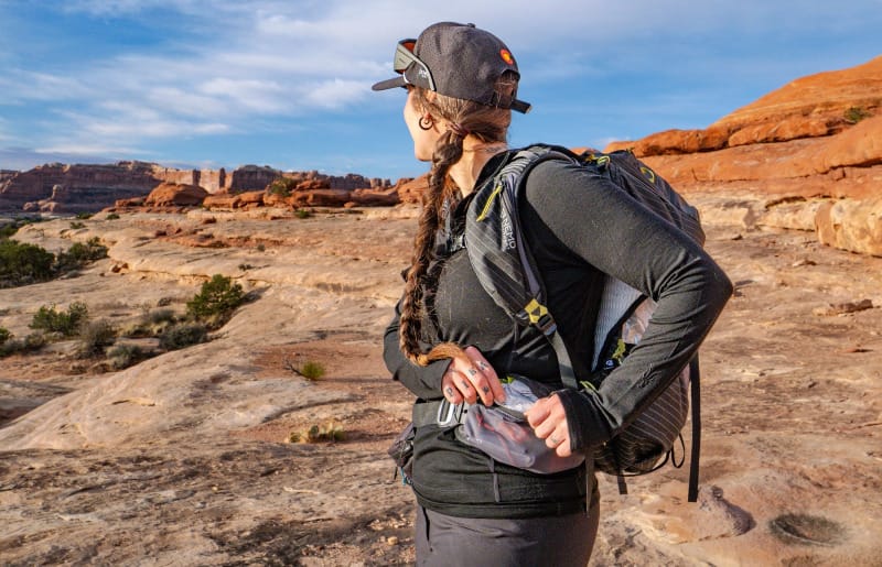 Hiker wearing a black sun hoodie and backpack looking across red rock formations and open slickrock terrain under a blue sky in the desert Southwest.