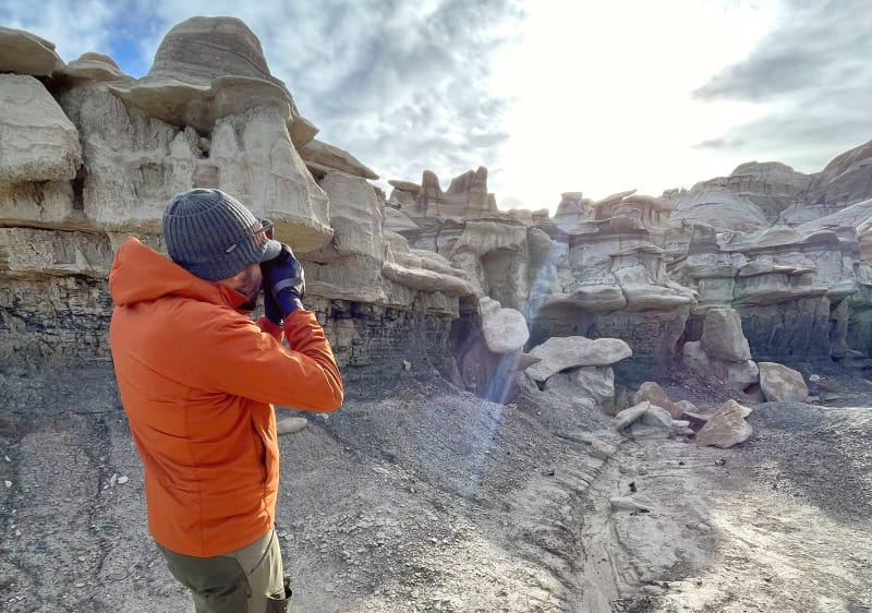 Hiker wearing an orange jacket photographing dramatic white rock hoodoos under a bright desert sky.