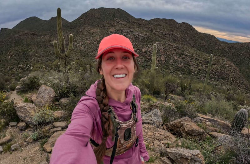 Hiker wearing a hooded sun shirt and red cap sitting among desert rocks with blooming wildflowers and saguaro cacti.