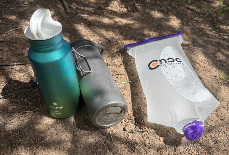 Three water containers—a Snow Peak bottle, a gray filter bottle, and a CNOC water bag—on forest ground in dappled sunlight.