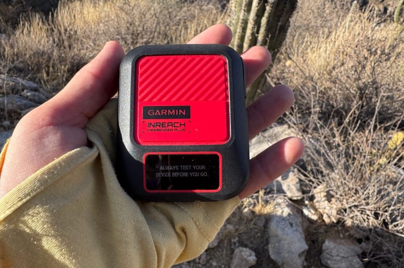 Hand holding a red Garmin inReach Messenger+ satellite communicator outdoors in a desert landscape with cactus and rocky terrain.