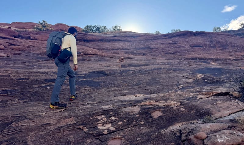 Hiker wearing a backpack climbing up a sandstone slope under a clear desert sky.