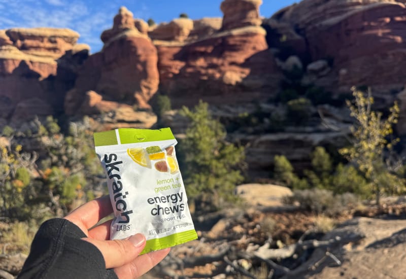 Close-up of a hand holding Skratch Labs lemon green tea energy chews with red rock formations and blue sky in the background.