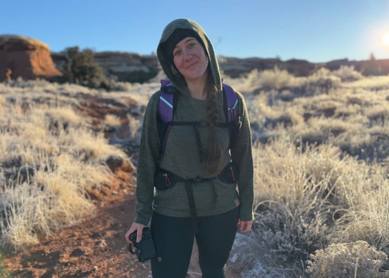 Hiker wearing a green alpaca wool PAKA sweater and backpack on a frosty desert trail at sunrise, holding a camera with red rock formations in the background.