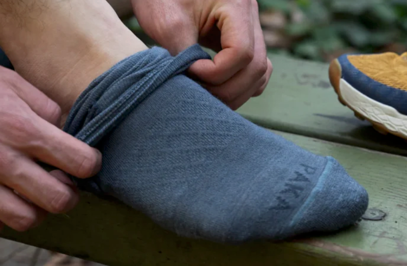 Hiker putting on blue PAKA alpaca socks beside trail shoes on a wooden bench.
