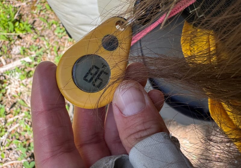 Close-up of a small yellow trail thermometer attached to a backpack with a hand holding it.