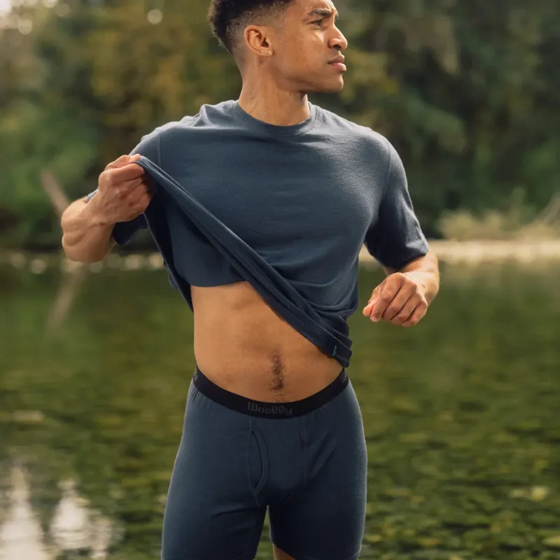 Man wearing Woolly Clothing merino T-shirt and boxer briefs beside a mountain lake, lifting his shirt to show breathable base layers.