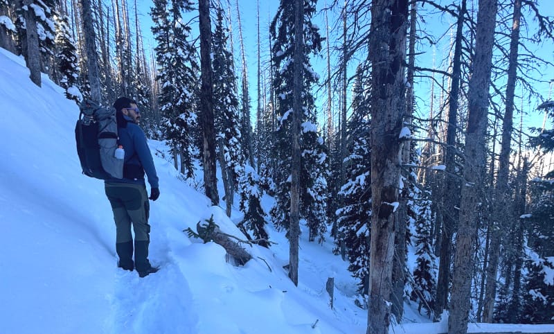 Side view of a hiker carrying the Outdoor Vitals Shadowlight Carbon 60 backpack on a narrow snowy mountain trail.