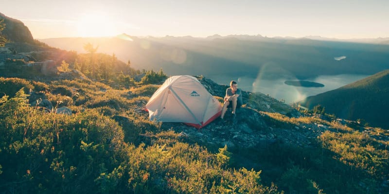 A person sitting beside an MSR tent on a mountaintop at sunrise, overlooking a sweeping landscape of mountains, forest, and lakes.