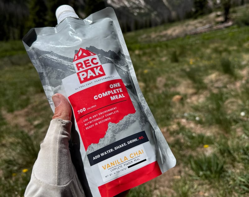 Hand holding a REPAK Vanilla Chai meal pouch with snow-capped mountains and pine forest in the background.