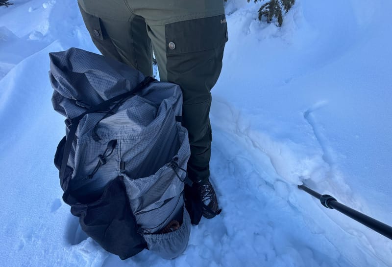Close-up of the Outdoor Vitals Shadowlight Carbon 60 backpack resting in deep snow, showing side pockets and durable UltraGrid material.