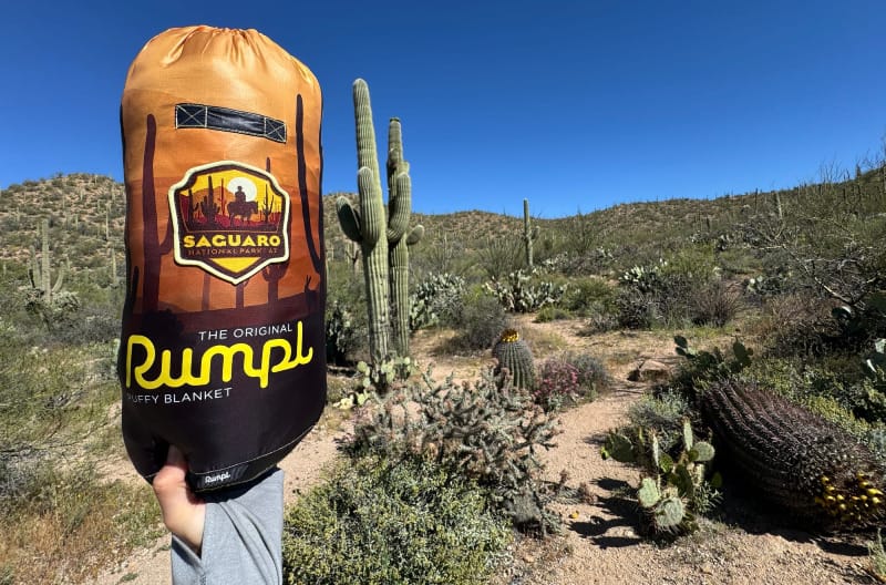 Hand holding a Rumpl Original Puffy Blanket in the Saguaro National Park edition with towering cacti and desert hills in the background under a bright blue sky.