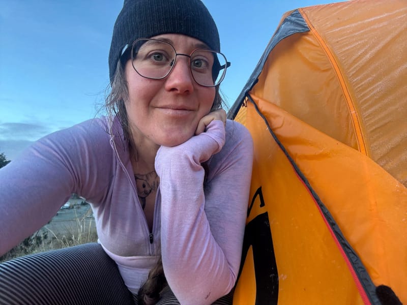 Woman taking a selfie beside an orange MSR tent at dawn, wearing glasses, a beanie, and a light purple base layer.