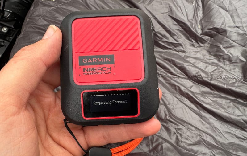 Hand holding a red Garmin inReach Messenger Plus SOS device on a tent floor, showing the small screen requesting a weather forecast.