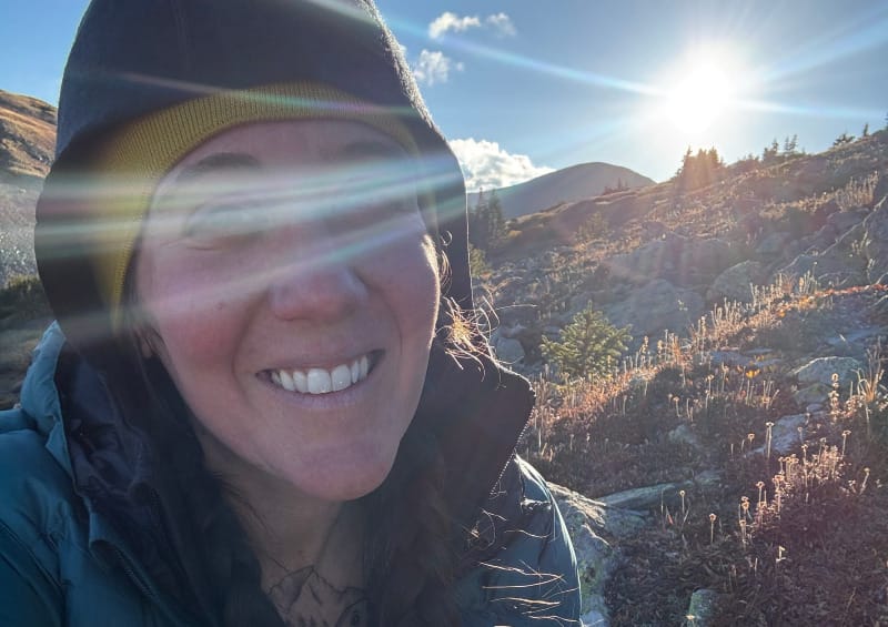 Close-up selfie of a hiker wearing the Ibex Mammoth Hoodie in the mountains at sunrise, with sun flares and frosty alpine terrain in the background.