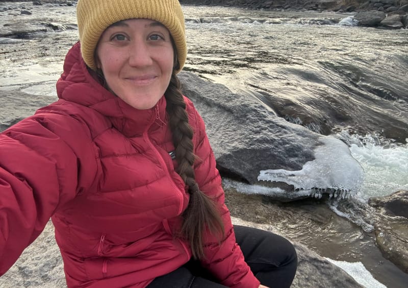 Hiker sitting beside a partially frozen river wearing a red Katabatic Gear Tincup down jacket and yellow beanie on a cold winter day.