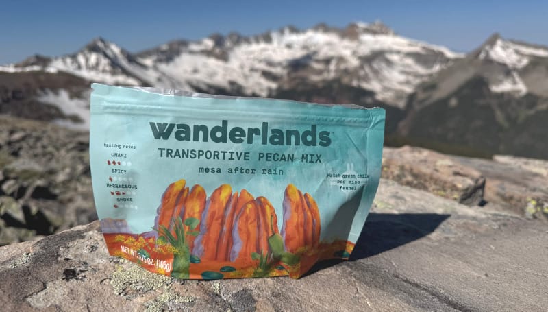 Bag of Wanderlands Transporitve Pecan Mix sitting on a rocky alpine ridge with snow-covered mountains and a bright blue sky in the background.